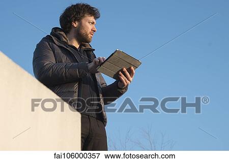 Stock Photo - Man holding digital tablet outdoors, looking away in thought. Fotosearch