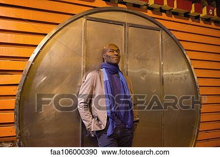 Stock Image - Man leaning against wall with hands in pockets. Fotosearch