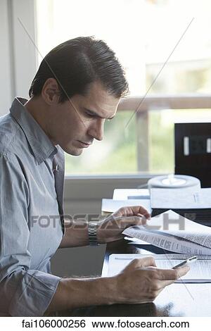 Stock Photograph - Man looking at smartphone while reading document. Fotosearch