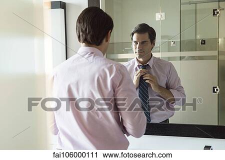 Stock Image - Man looking in bathroom mirror, adjusting necktie. Fotosearch
