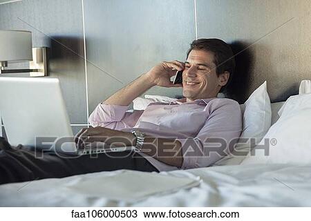 Stock Image - Man lying in bed, using cell phone and laptop computer. Fotosearch