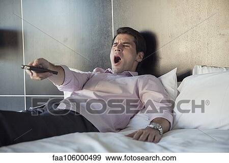 Stock Photo - Man lying on bed, yawning while watching TV. Fotosearch