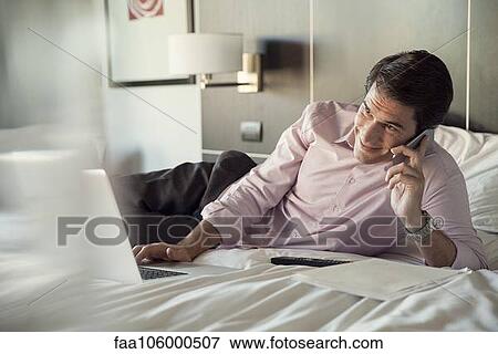 Man lying on hotel bed, talking on cell phone and using laptop computer View Large Photo Image Stock Photo - Man lying on hotel bed, talking on cell phone and using laptop computer. Fotosearch