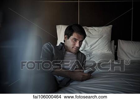 Picture - Man relaxing on bed with smartphone. Fotosearch
