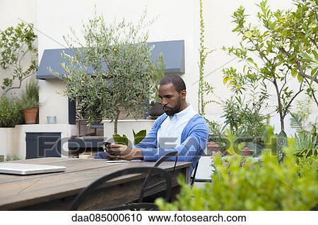 Man seated at table in hotel courtyard using cell phone View Large Photo Image Stock Image - Man seated at table in hotel courtyard using cell phone. Fotosearch