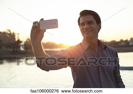 Man using smartphone to photograph himself in front of sunset View Large Photo Image Stock Photograph - Man using smartphone to photograph himself in front of sunset. Fotosearch