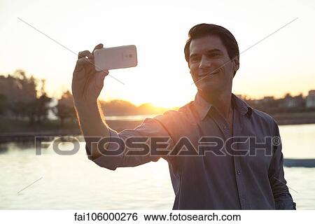 Man using smartphone to photograph himself in front of sunset View Large Photo Image Stock Photograph - Man using smartphone to photograph himself in front of sunset. Fotosearch