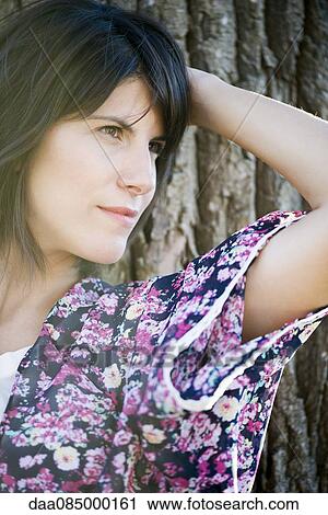 Stock Image - Woman leaning against tree trunk, looking away in thought. Fotosearch