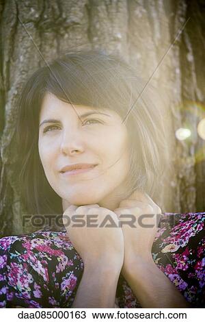 Stock Image - Woman leaning against tree trunk, smiling, portrait. Fotosearch