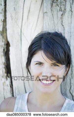 Stock Photo - Woman leaning against tree trunk, smiling cheerfully, portrait. Fotosearch