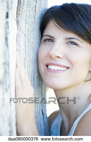 Stock Photograph - Woman leaning against tree trunk, smiling cheerfully. Fotosearch