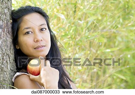 Stock Image - Woman leaning against tree trunk with apple in hand, portrait. Fotosearch