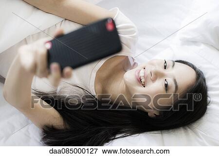 Woman lying in bed, using smartphone to take a selfie View Large Photo Image Stock Photo - Woman lying in bed, using smartphone to take a selfie. Fotosearch