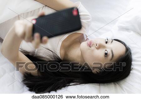 Woman lying in bed, using smartphone to take a selfie View Large Photo Image Stock Photograph - Woman lying in bed, using smartphone to take a selfie. Fotosearch