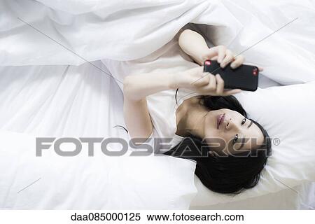 Stock Photography - Woman lying in bed, using smartphone to take a selfie. Fotosearch