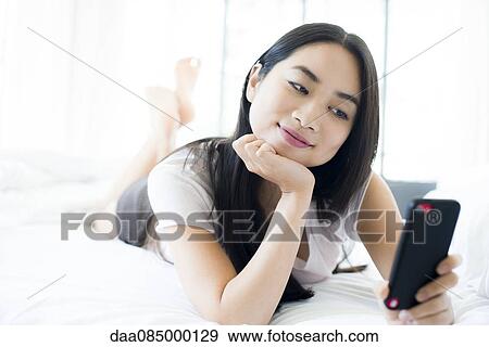 Woman lying on bed, smiling at smartphone View Large Photo Image Stock Photo - Woman lying on bed, smiling at smartphone. Fotosearch