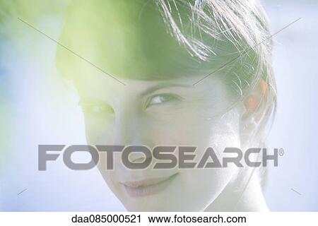 Stock Image - Woman smiling through foliage, portrait. Fotosearch