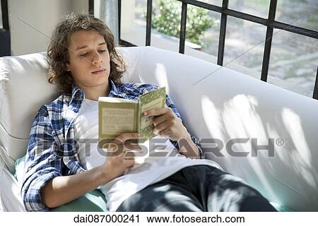 Stock Image - Young man reading book on couch. Fotosearch