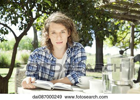 Young man sitting at breakfast table outdoors with book View Large Photo Image Stock Photo - Young man sitting at breakfast table outdoors with book. Fotosearch