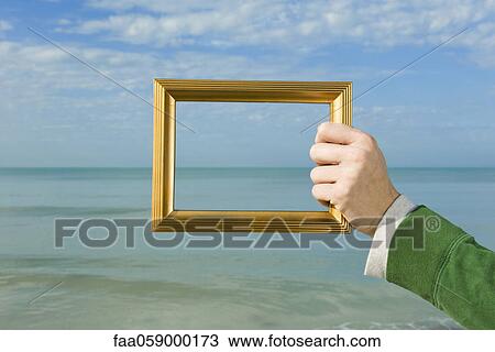 Stock Image - Person holding picture frame in front of ocean. Fotosearch