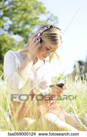 Young woman sitting in tall grass listening to headphones View Large Photo Image Picture - Young woman sitting in tall grass listening to headphones. Fotosearch