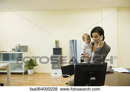 Stock Photo - Mother holding baby while taking phone call in office. Fotosearch