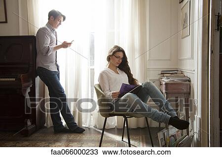 Stock Image - Couple relaxing at home, woman reading magazine, man looking at cell phone. Fotosearch