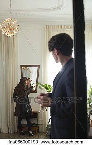Stock Image - Man waiting impatiently in doorway while woman applies lipstick in mirror. Fotosearch