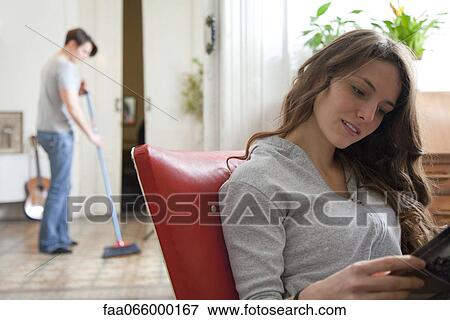 Stock Photo - Woman reading magazine while her husband sweeps the floor. Fotosearch