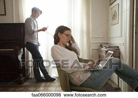 Woman using laptop computer at home, man holding cell phone in background View Large Photo Image Stock Photo - Woman using laptop computer at home, man holding cell phone in background. Fotosearch