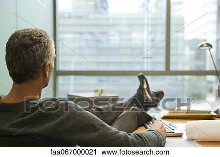 Stock Image - Executive sitting in office with feet on desk, rear view. Fotosearch