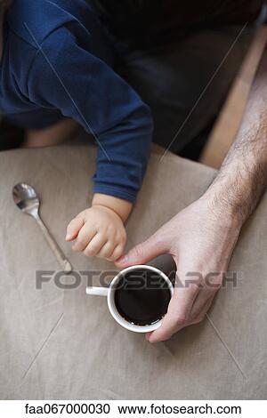 Stock Image - Man having cup of coffee while holding toddler son, cropped. Fotosearch
