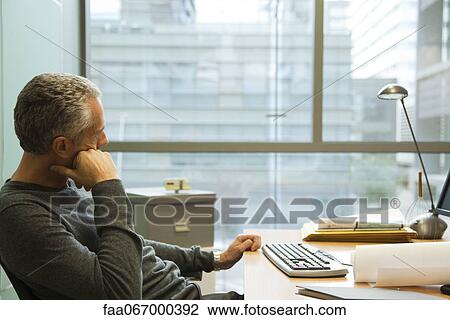 Stock Image - Man sitting at desk in office, looking away in thought. Fotosearch