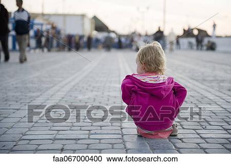 Stock Image - Toddler girl sitting on the ground in city square, watching people walk by. Fotosearch