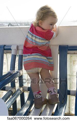 Toddler girl standing on chair, peeking over balcony railing, rear view View Large Photo Image Picture - Toddler girl standing on chair, peeking over balcony railing, rear view. Fotosearch