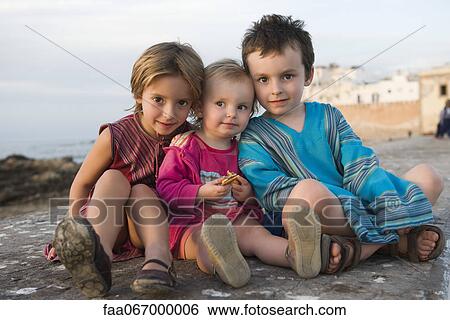Stock Photograph - Young siblings sitting together on rocky beach, portrait. Fotosearch