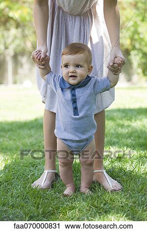 Stock Image - Baby standing outdoors, holding mother's hands for support. Fotosearch