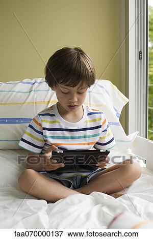 Boy sitting on bed, reading e-book View Large Photo Image Stock Photo - Boy sitting on bed, reading e-book. Fotosearch