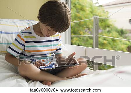 Boy sitting on bed, using digital tablet View Large Photo Image Picture - Boy sitting on bed, using digital tablet. Fotosearch