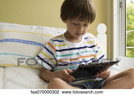 Boy sitting on bed, using digital tablet View Large Photo Image Stock Image - Boy sitting on bed, using digital tablet. Fotosearch