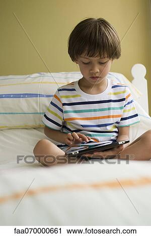 Boy sitting on bed, using digital tablet View Large Photo Image Stock Image - Boy sitting on bed, using digital tablet. Fotosearch
