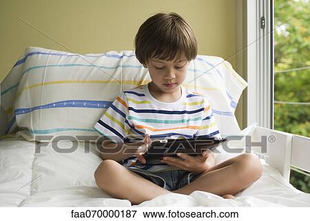 Boy sitting on bed, using digital tablet View Large Photo Image Stock Photo - Boy sitting on bed, using digital tablet. Fotosearch