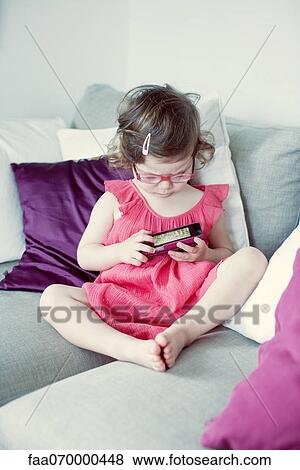 Little girl sitting on couch looking at picture on cell phone View Large Photo Image Stock Photo - Little girl sitting on couch looking at picture on cell phone. Fotosearch