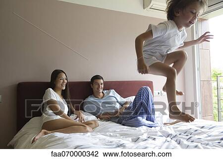 Stock Image - Parents watching as young son jumps on bed. Fotosearch