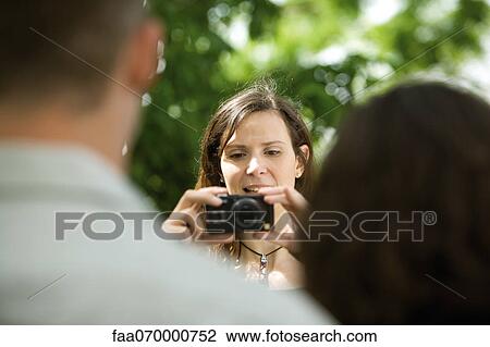 Stock Image - Woman photographing couple with digital camera, over the shoulder view. Fotosearch