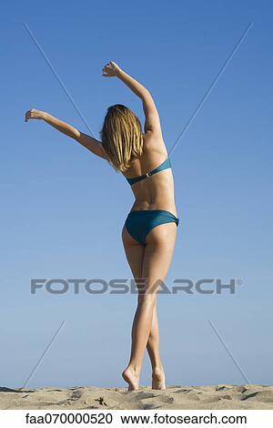 Stock Image - Woman walking on beach with arms raised in air, rear view. Fotosearch