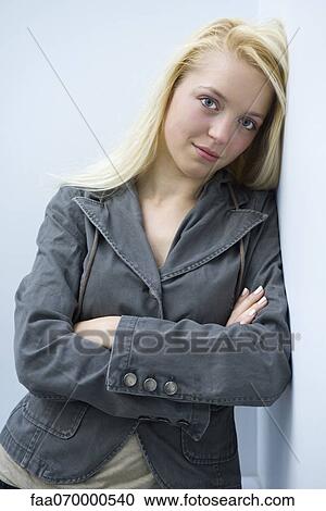 Stock Image - Young woman leaning against wall with arms folded, portrait. Fotosearch