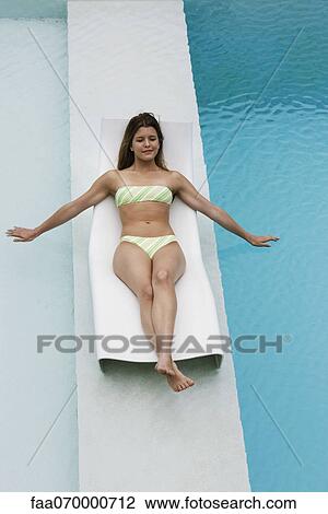 Stock Image - Young woman reclining on deck chair on walkway between swimming pools. Fotosearch