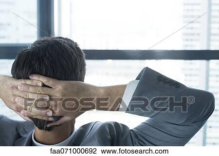 Stock Image - Businessman looking through window with hands behind head, rear view. Fotosearch