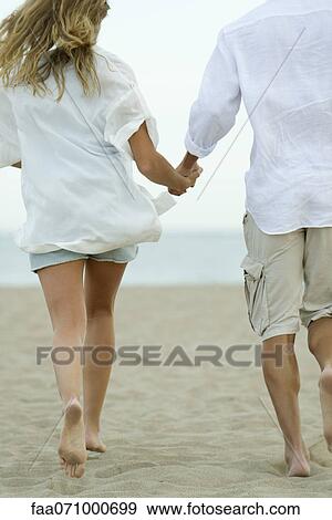 Stock Photo - Couple running hand in hand at the beach, rear view. Fotosearch